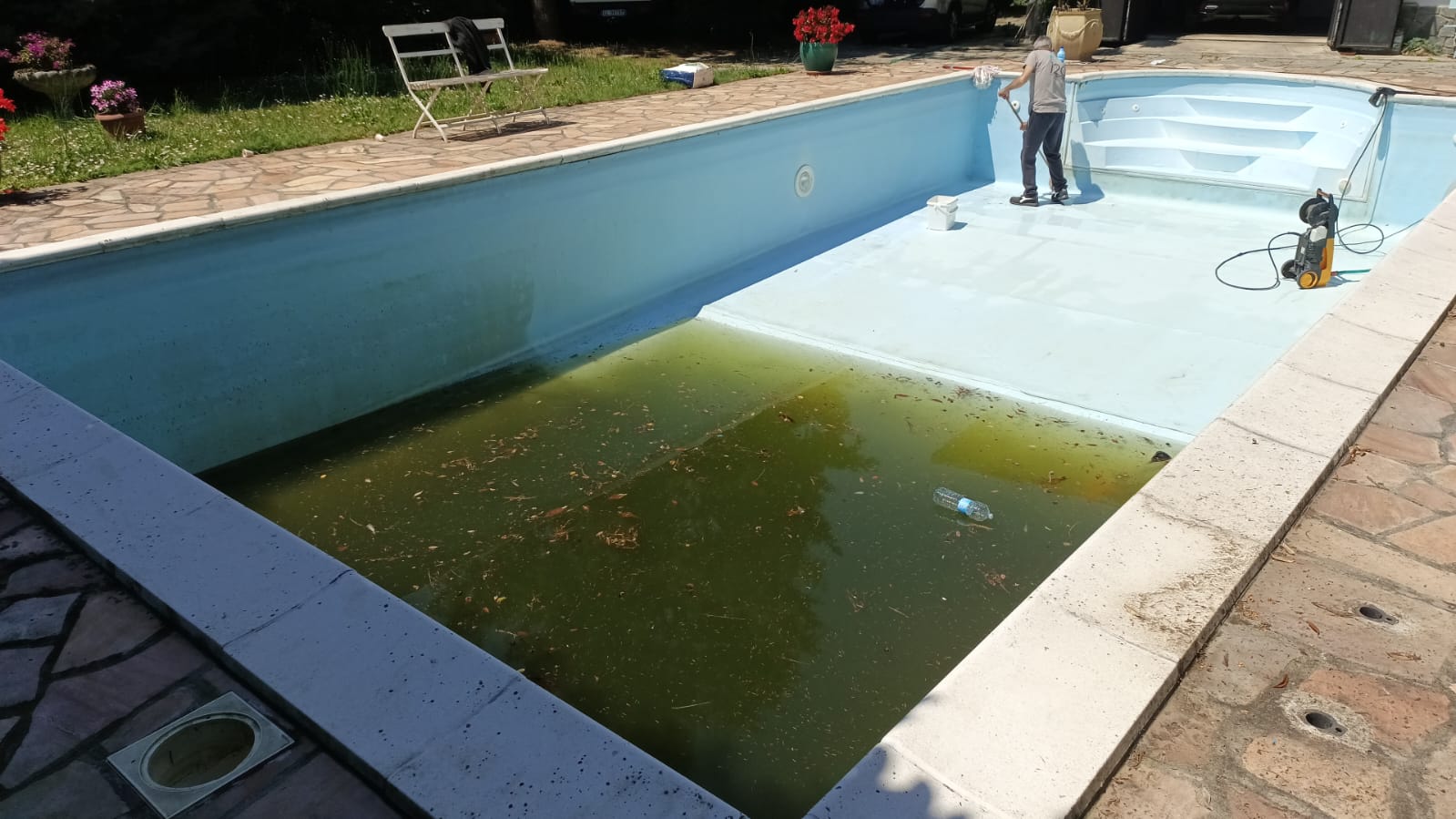 Piscina azzurra in giardino, con acqua verde sporca, un uomo sul bordo e attrezzi per la pulizia.